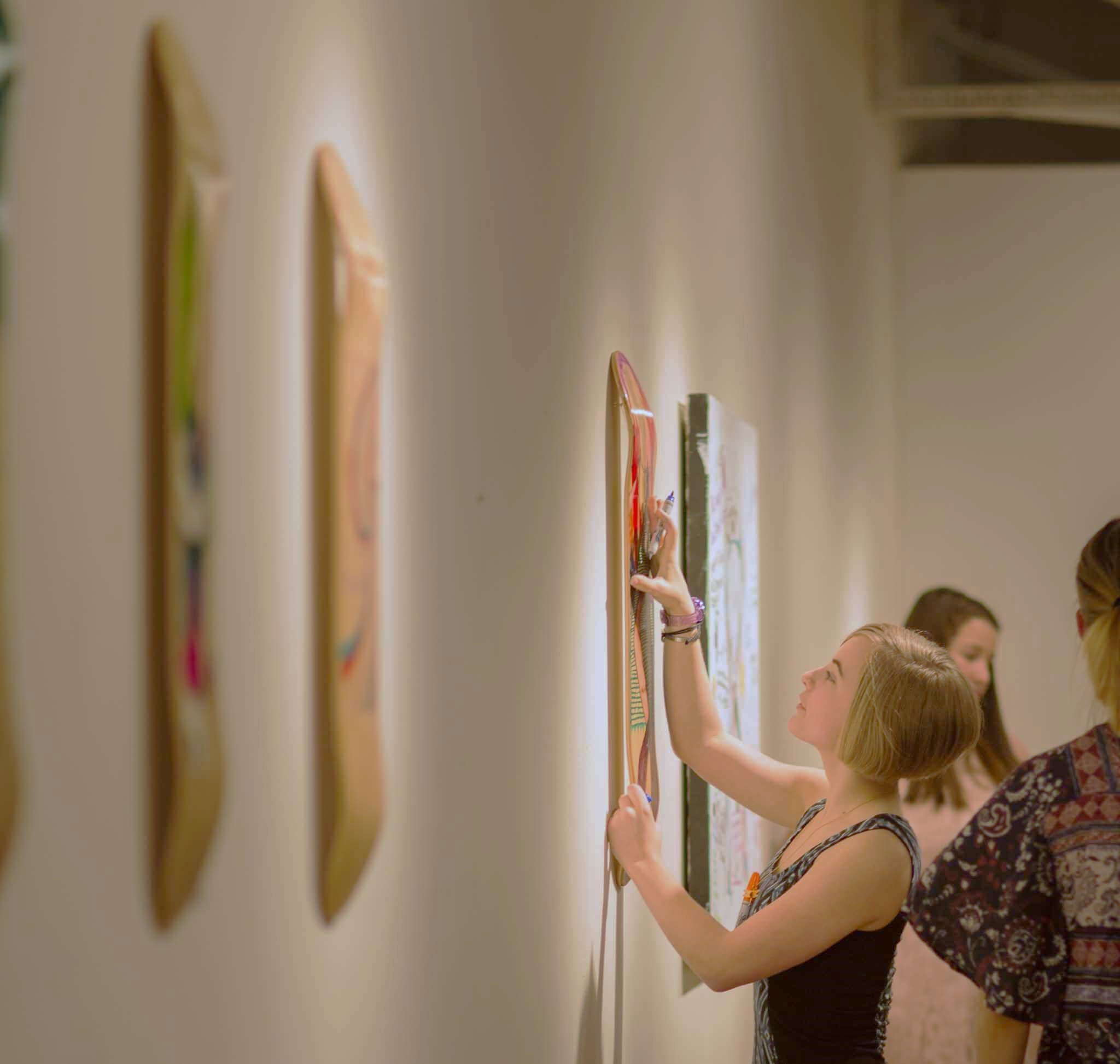 Blonde woman touching a skateboard on a wall in an art gallery..