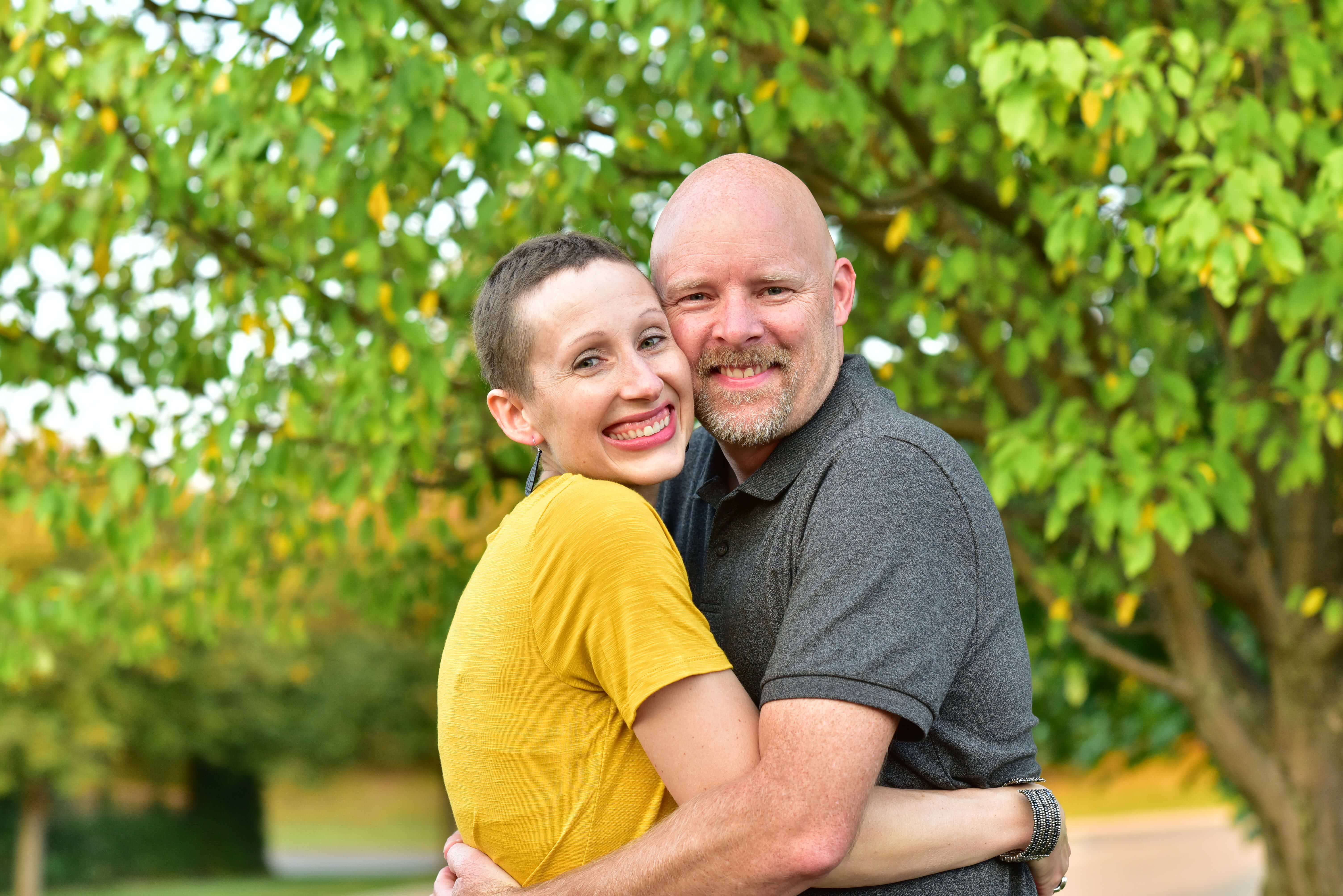 A smiling couple hugs against a leafy background.